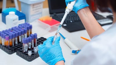 A close-up, over-the-shoulder shot shows a scientist, transferring liquid from one test tube to another using a pipette
