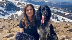 A woman sits on a mountainside with her dog by her side